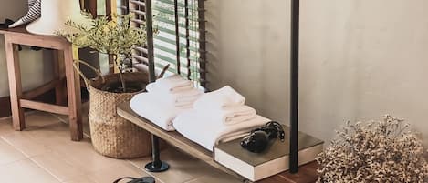 A cozy room with a bed, a small table with towels, a chair, a window with blinds, and a basket with a book.