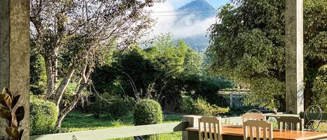 A covered outdoor seating area with a table and chairs, overlooking a lush garden and a mountain in the distance.