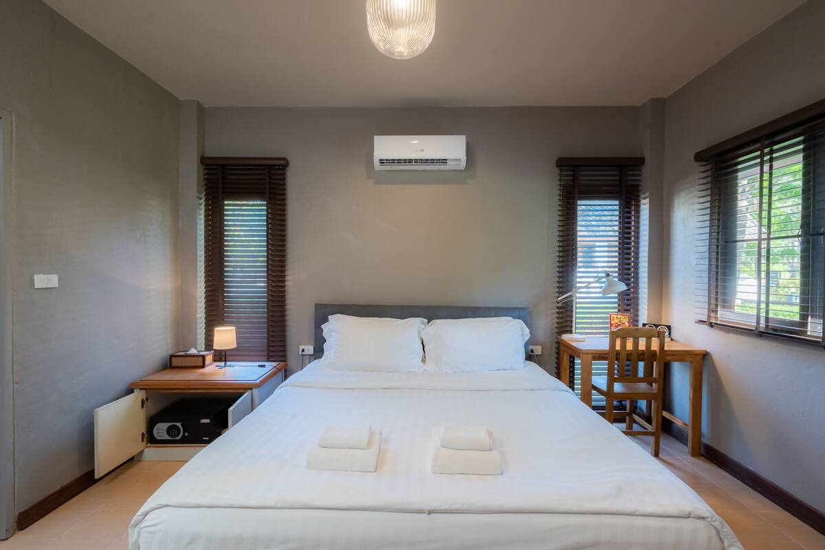 A hotel room with a large bed, a desk, a chair, an air conditioner, and a window with blinds.