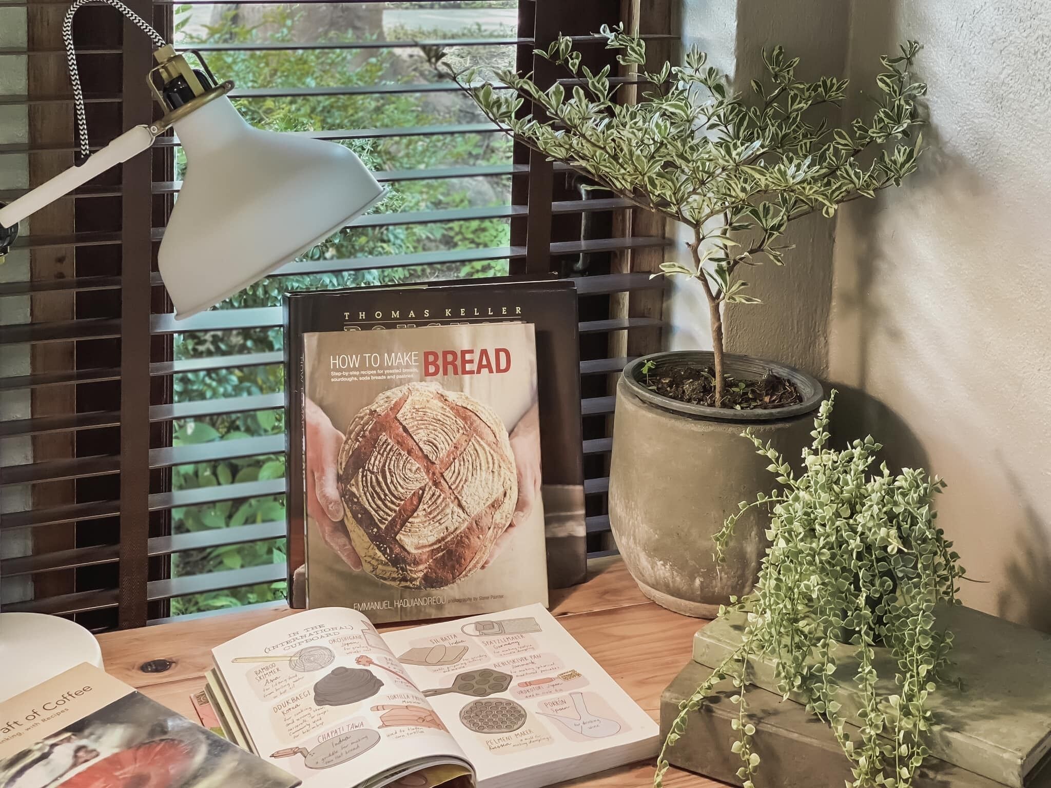 Un libro titulado 'How to Make Bread' de Thomas Keller, una planta en maceta y una lámpara sobre una mesa junto a una ventana con persianas.