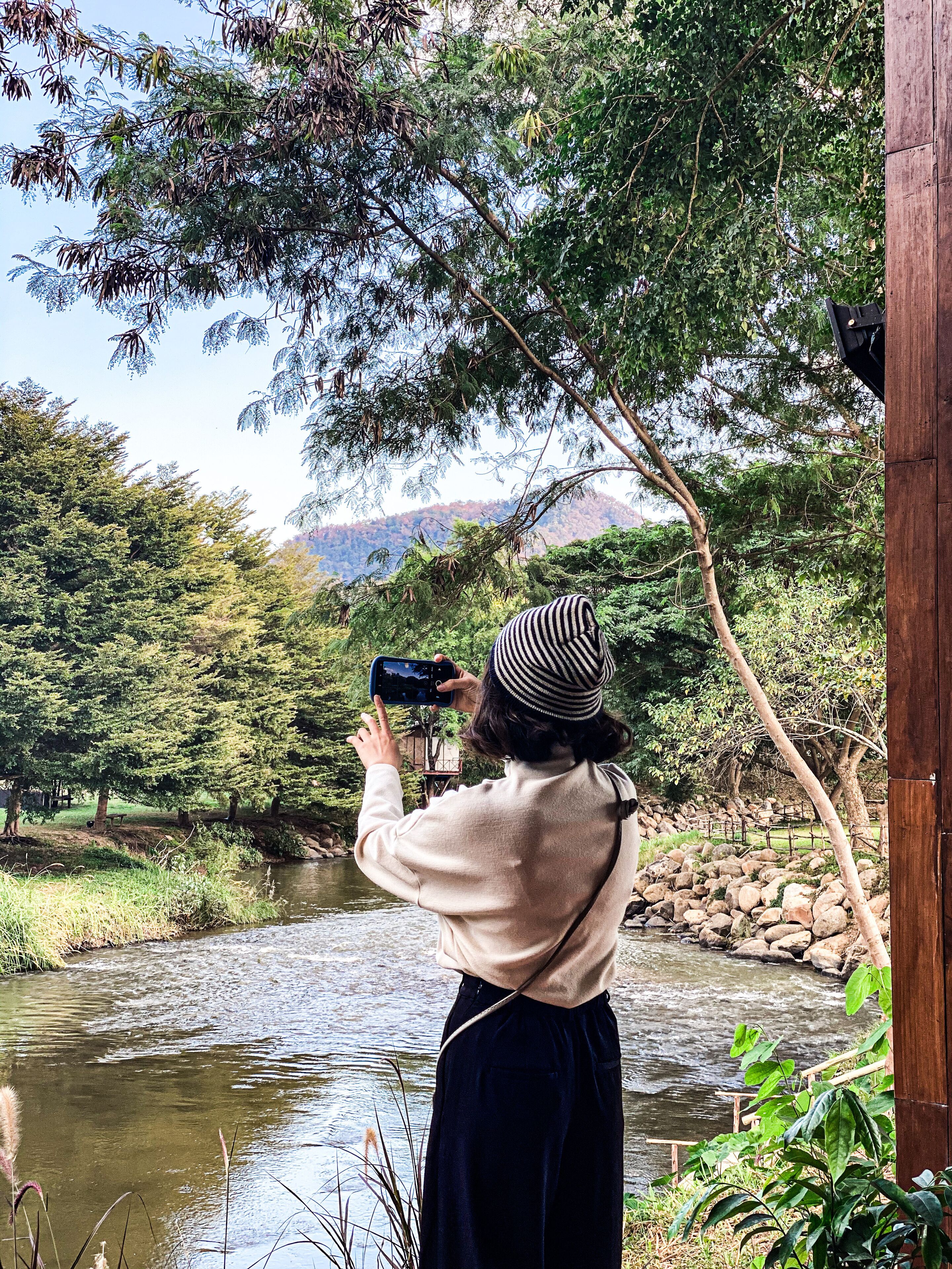 Una persona fotografiando un río con árboles y una estructura de madera al fondo.
