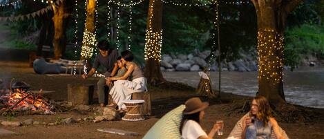 A campsite with a fire, people sitting on benches and bean bags, and trees decorated with lights.