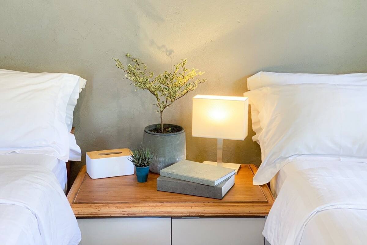 A neatly made bed with white linens, a bedside table with a lamp, a potted plant, and a small box.