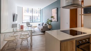 Cozinha moderna com ilha central, área de jantar com mesa de vidro e cadeiras de metal, e sala de estar com sofá e planta em vaso.