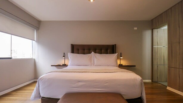 A modern bedroom with a large bed, bedside tables, and a view of the city through the window. - w * | Elegant 1BR in San Isidro (San Isidro)