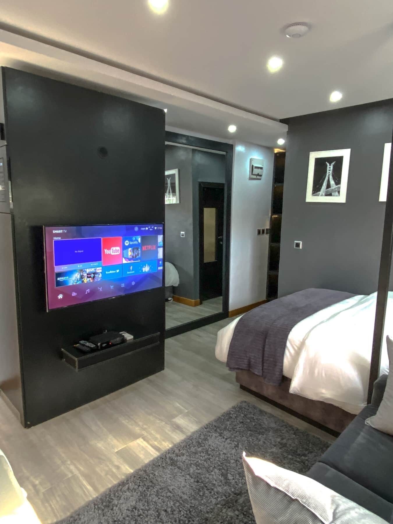 A modern bedroom with a flat-screen TV mounted on a black wall, a bed with a gray blanket, a gray carpet, and a mirror reflecting the room.