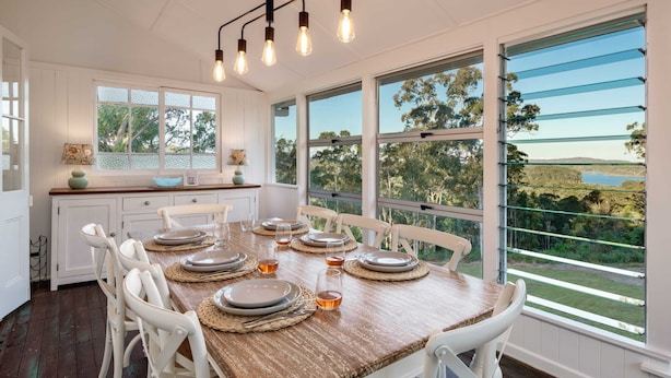 A dining area with a wooden table set for six, white chairs, and large windows offering a view of greenery and a lake. - Lakeview Cottage in Mooloolah Valley: Pool & Sauna (Mooloolah Valley)