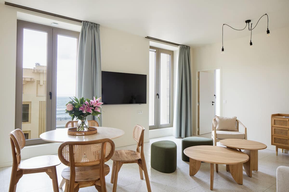 Un salon moderne avec une table ronde en bois, deux chaises en bois, un pouf vert et un canapé beige. Un téléviseur à écran plat est fixé au mur, et il y a une fenêtre avec des rideaux.
