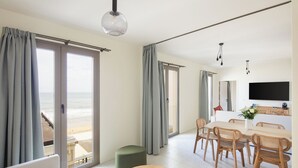 A modern living room with a large window overlooking the beach, a dining area with wooden furniture, and a flat-screen TV mounted on the wall. - Hôtel La Suite (Biarritz)