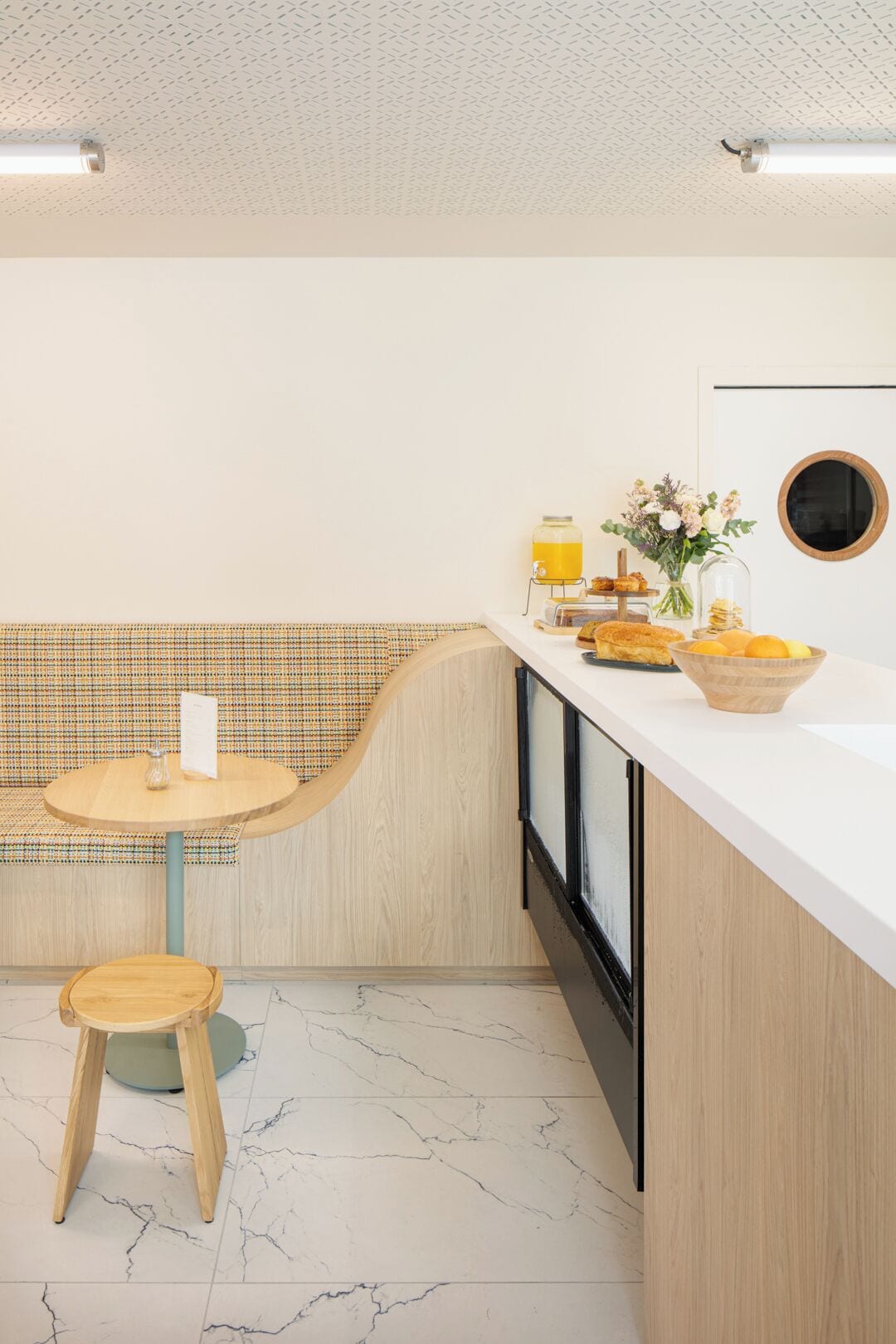 A modern kitchen with a wooden bench, a round table, and a breakfast setup including a fruit bowl and a vase with flowers.