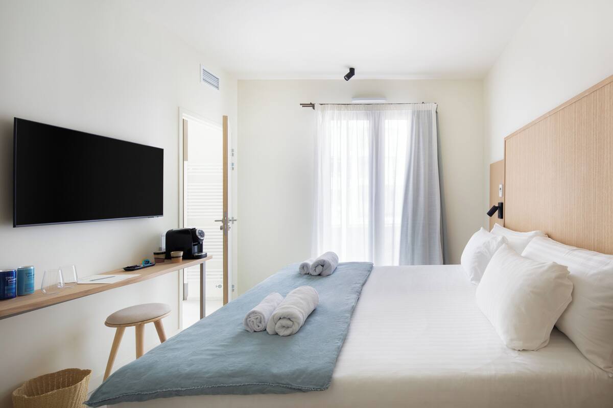 Une chambre d’hôtel avec un lit, un téléviseur fixé au mur, une petite table avec une machine à café et une fenêtre avec des rideaux.