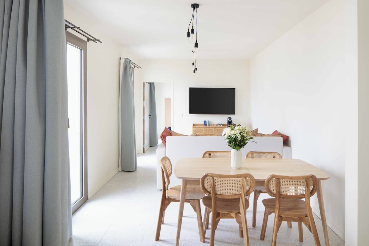 Un espace repas moderne avec une table et des chaises en bois, un téléviseur à écran plat fixé au mur et une grande fenêtre avec des rideaux.
