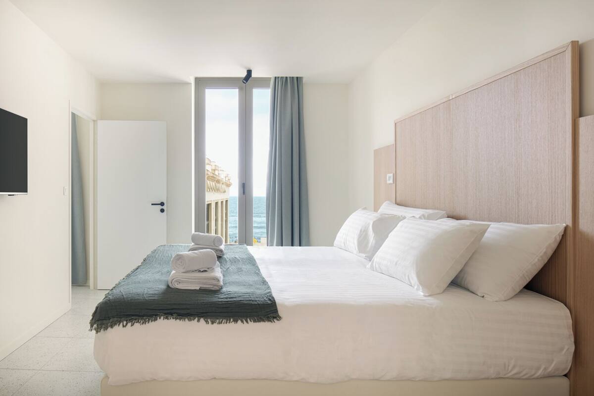 Une chambre d’hôtel avec un lit, une télévision, un balcon avec des rideaux et une vue sur la mer.