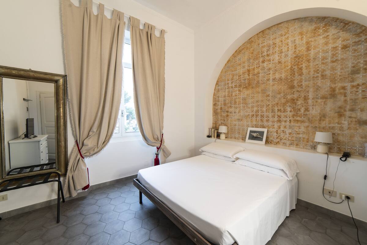 Un dormitorio con cama, espejo, ventana con cortinas, lámpara de pared y pared de azulejos.