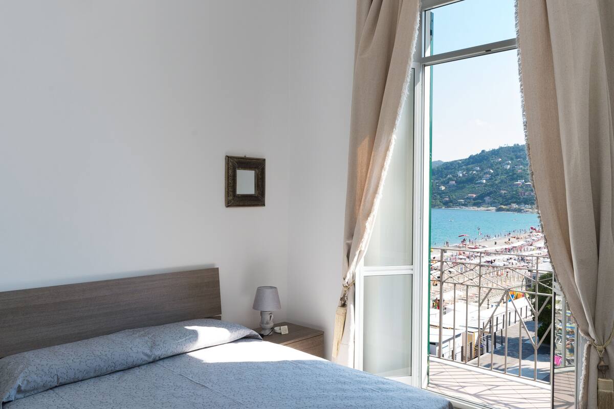 Un dormitorio con cama, mesita de noche, lámpara y vista a la playa a través de un ventanal amplio.