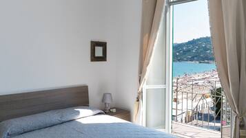 Une chambre à coucher avec un lit, une table de chevet, une lampe et une vue sur la plage, visible à travers une grande fenêtre.