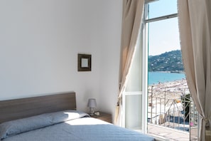 A bedroom with a bed, a nightstand, a lamp, and a view of a beach through a large window. - Hotel Miramare - Spotorno (Spotorno)