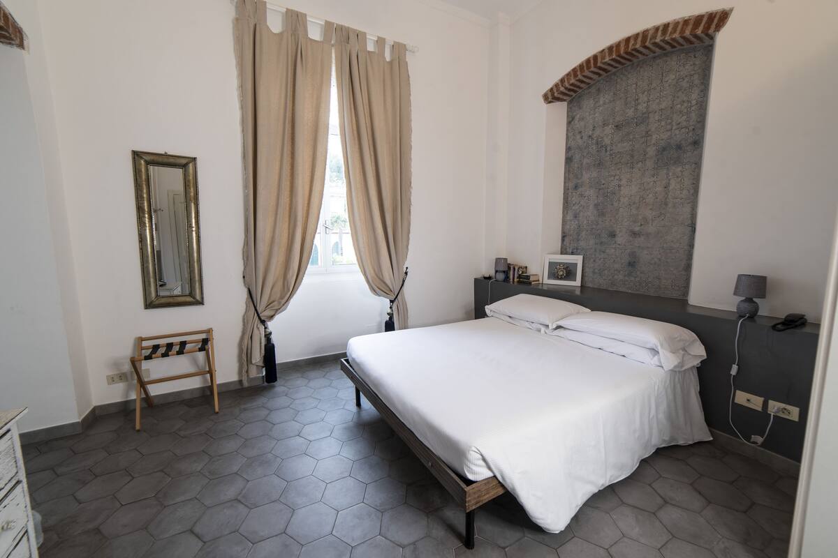 Un dormitorio con cama, cortinas, un espejo, una mesita de noche y una lámpara de pared.