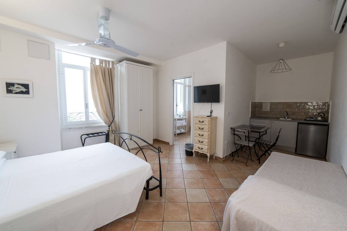 Habitación de hotel con dos camas, kitchenette, televisor y balcón con cortinas.