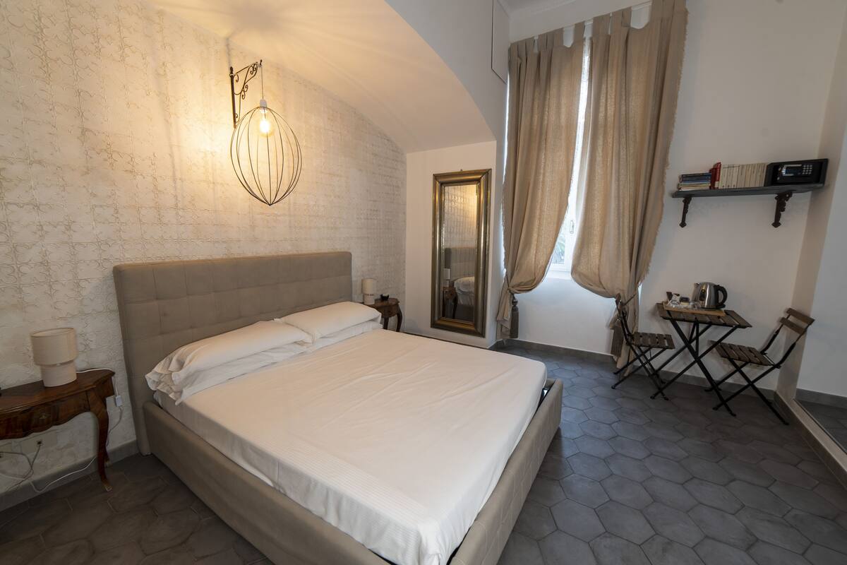 Un dormitorio con una cama grande, una mesita de noche con lámpara, una pequeña mesa con hervidor y una silla.