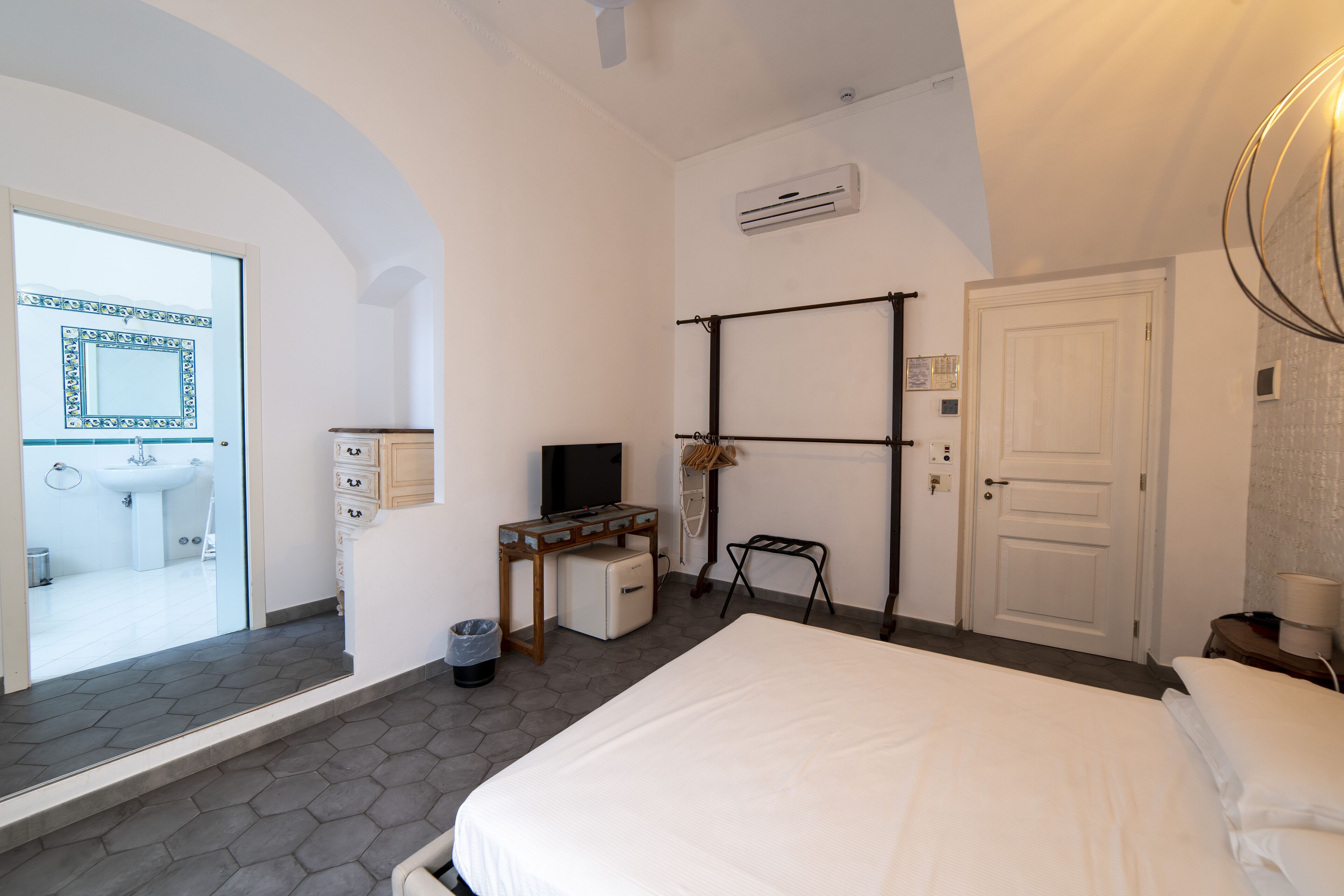 A hotel room with a large bed, a desk with a computer, a TV, and a bathroom visible through an arched doorway.