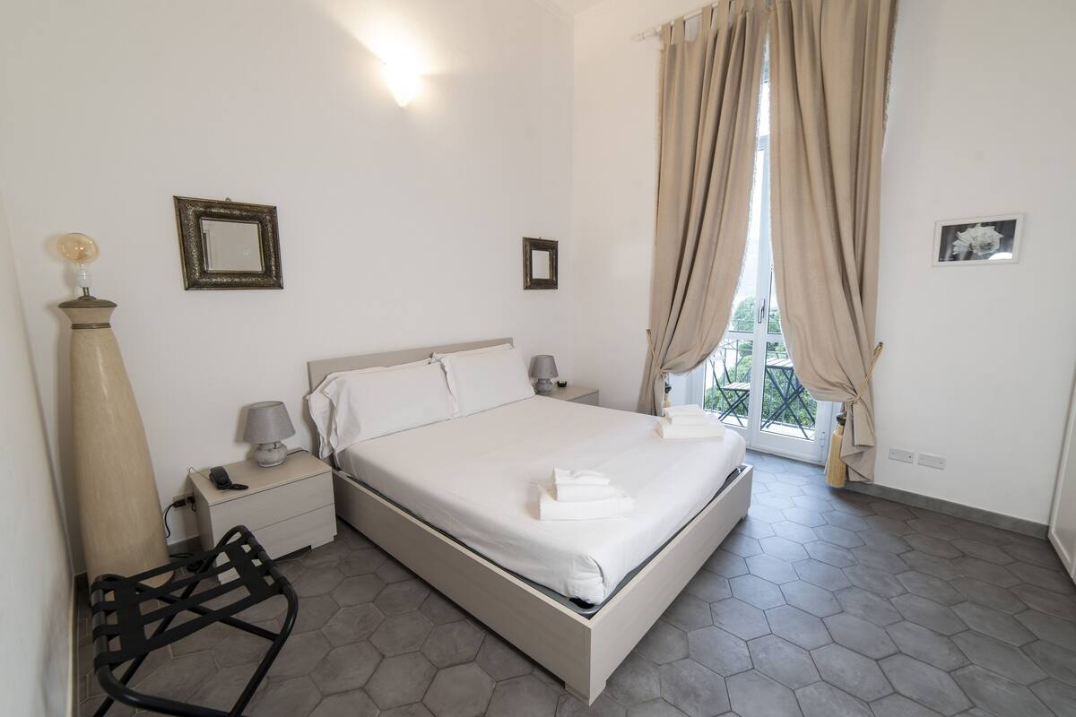 Una habitación de hotel con cama, mesitas de noche, lámpara, silla y ventana con cortinas.