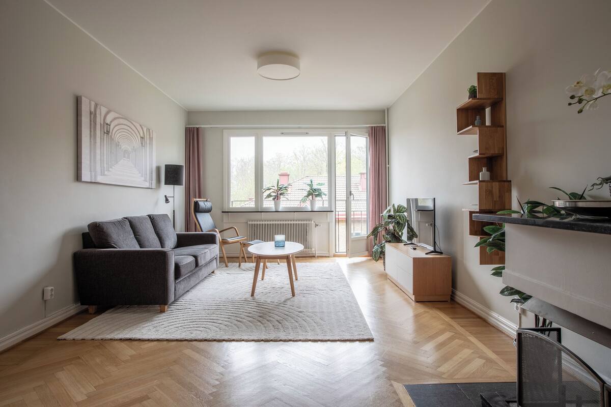 A modern living room with a grey sofa, a coffee table, and a TV. There is a large window with curtains, a radiator, and a plant on the windowsill.
