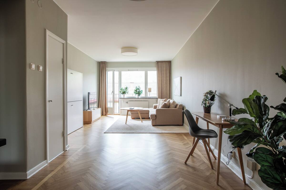 A modern living room with a sofa, coffee table, and a dining area with a table and chairs. There is a large window with curtains, a radiator, and a plant.