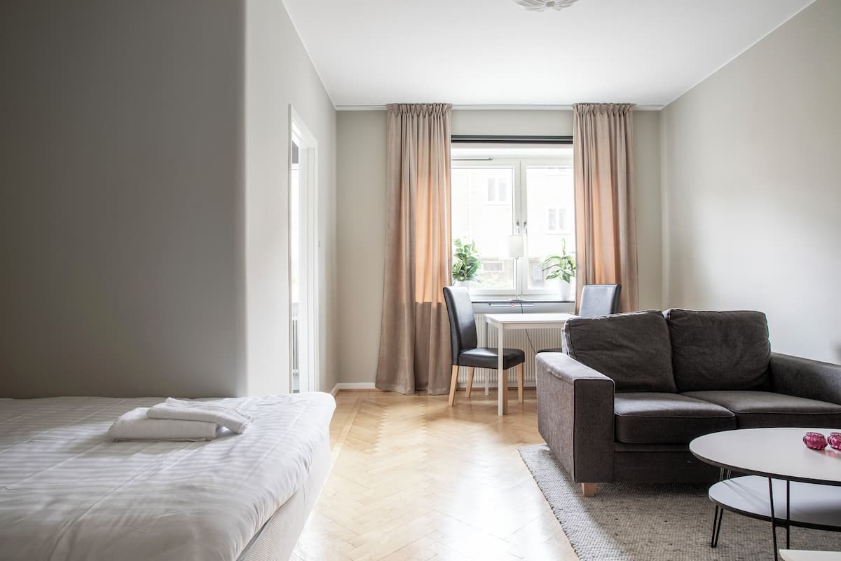 A modern hotel room with a bed, a sofa, a small table, and a window with curtains.