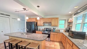 A modern kitchen with granite countertops, wooden cabinets, a black refrigerator, a stove, and a sink. There are three bar stools and a window providing natural light.