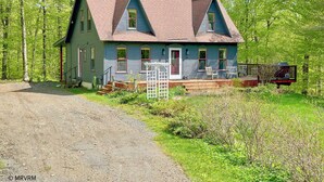 A two-story house with a steeply pitched roof, a front porch with a white railing, and a small deck area.