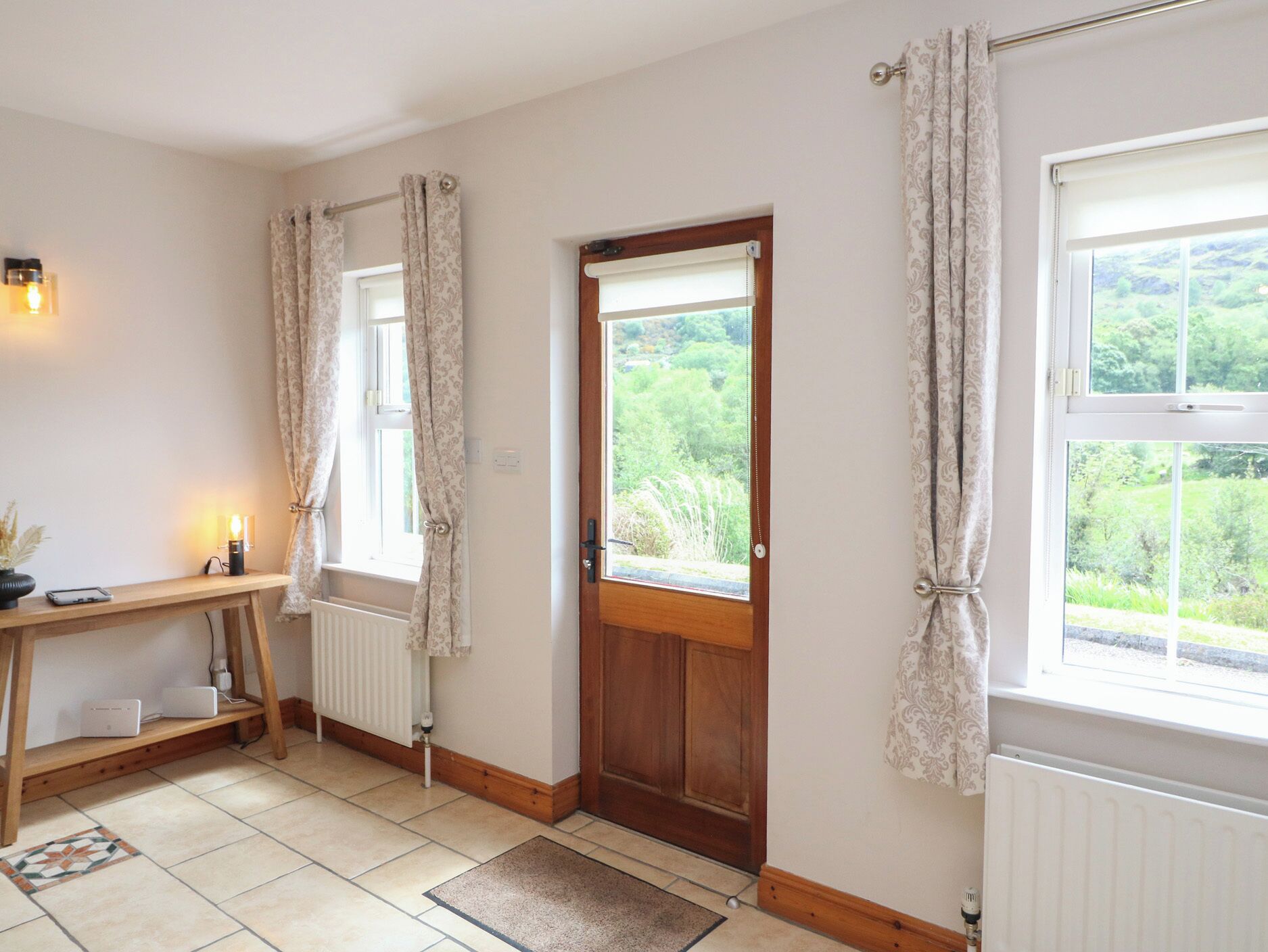 A room with a wooden door, a window with curtains, a radiator, and a small table with a lamp.