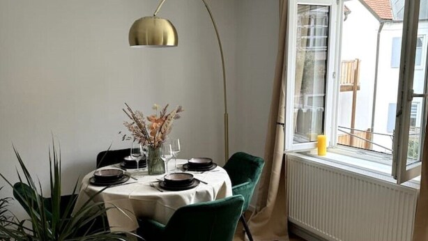 Un coin repas avec une table ronde dressée pour quatre personnes, des chaises vertes, un vase avec des fleurs séchées et un luminaire suspendu doré.