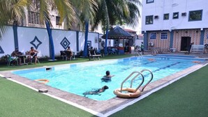 A swimming pool with two people swimming, surrounded by green artificial turf, palm trees, and a building with a tiled facade. - De Primtex HOtel Portharcourt (Port Harcourt)