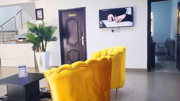 A reception area with a dark wooden door, a television mounted on the wall, and a pair of yellow chairs. - De Primtex HOtel Portharcourt (Port Harcourt)