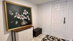 A hallway with a large framed painting, a white door, a black bench, and a patterned rug. - The Boho Oasis: Spacious Deluxe Apartment, Close to All Amenities, Safe & Quiet (Windsor)