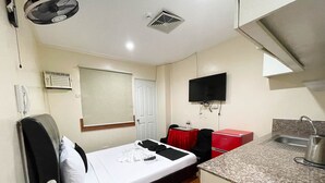 Een compacte hotelkamer met een enkel bed, een kleine kitchenette, een aan de muur bevestigde televisie en een raam met een zonwering.