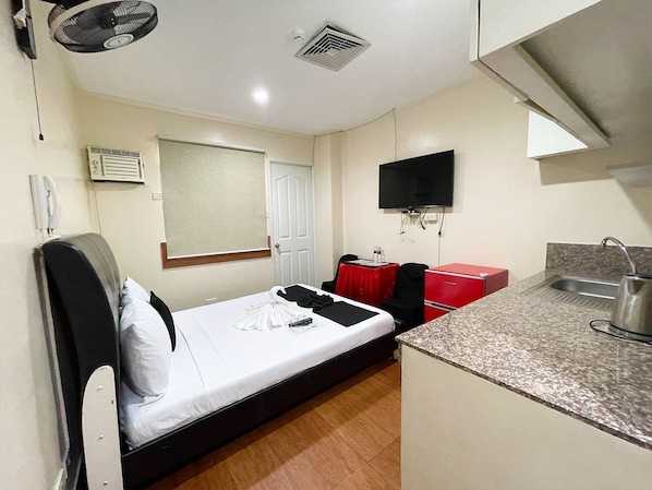 Habitación de hotel pequeña con una cama individual, una kitchenette compacta, un televisor montado en la pared y una ventana con persiana.