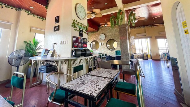 Een restaurant met een hoge plafond, houten balken en een centrale kolom met een klok. Er zijn meerdere tafels en stoelen, en de muren zijn versierd met spiegels en klokken.