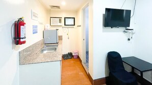 Een compacte kitchenette met een spoelbak, een kleine koelkast en een brandblusser. Er is een televisie aan de muur bevestigd en een stoel in de hoek.