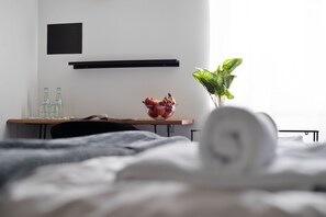 Un lit bien fait, avec une serviette roulée, un bol de fruits et une plante en pot sur une table de chevet.
