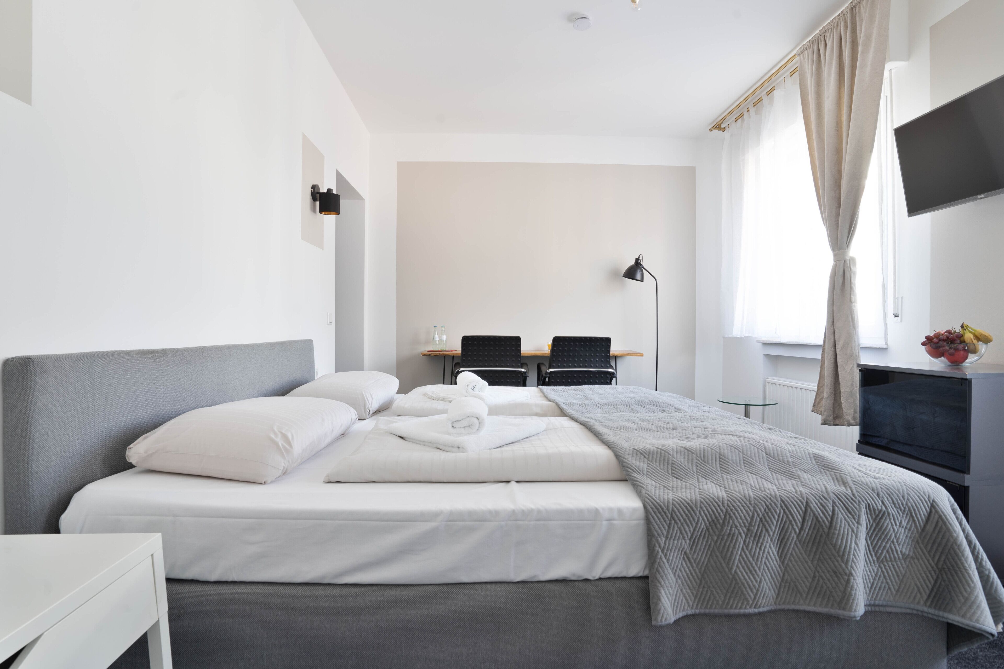 A modern hotel room with a large bed, a desk, two chairs, a flat-screen TV, and a window with curtains.