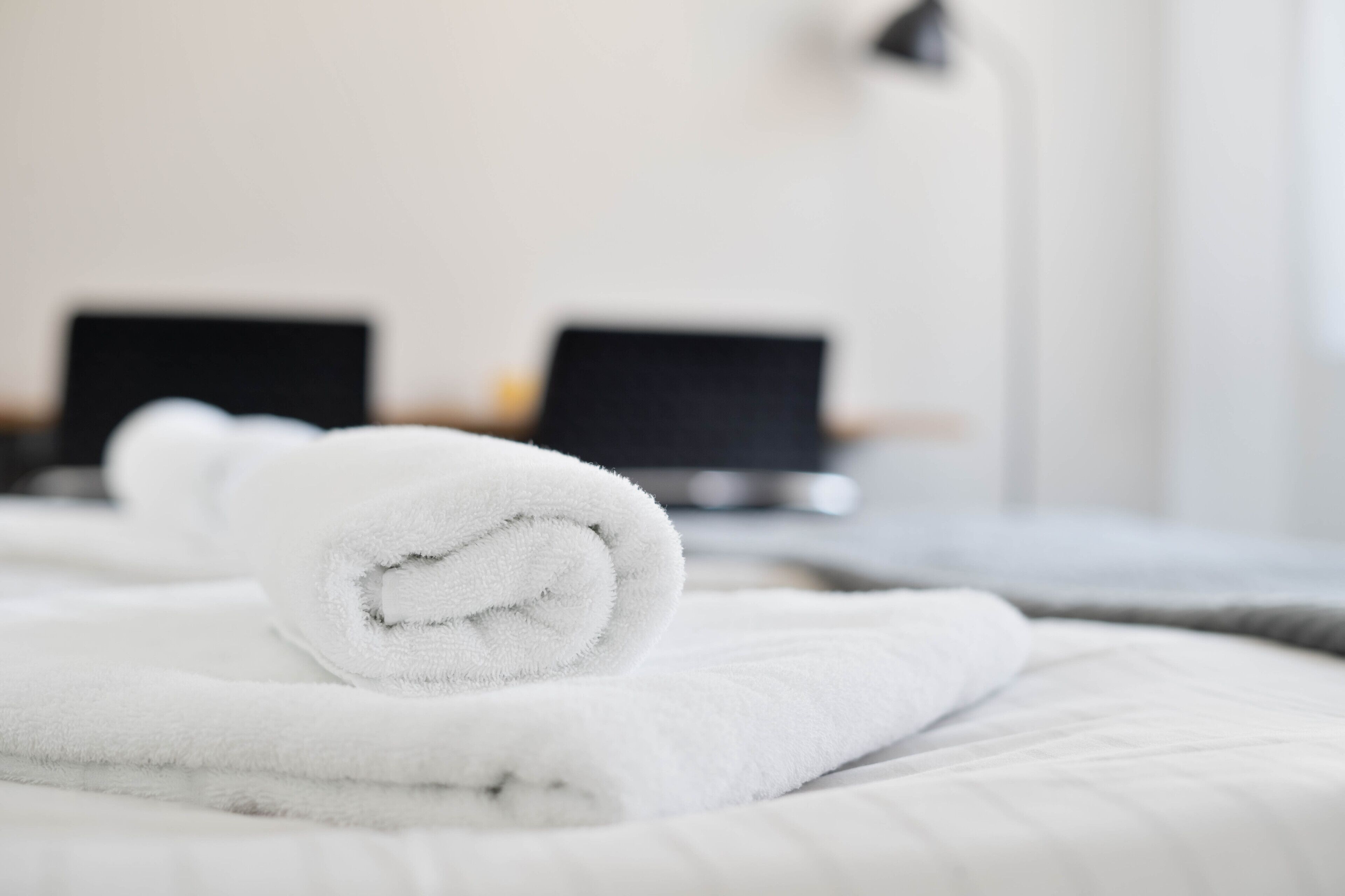 A neatly made bed with a rolled white towel in the foreground and two laptops on the nightstands in the background.