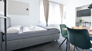 A modern hotel room with a grey sofa bed, a dining table with chairs, a flat-screen TV, and a large window with curtains. - Stilvolle Apartments in Bonn I home2share (Bonn)