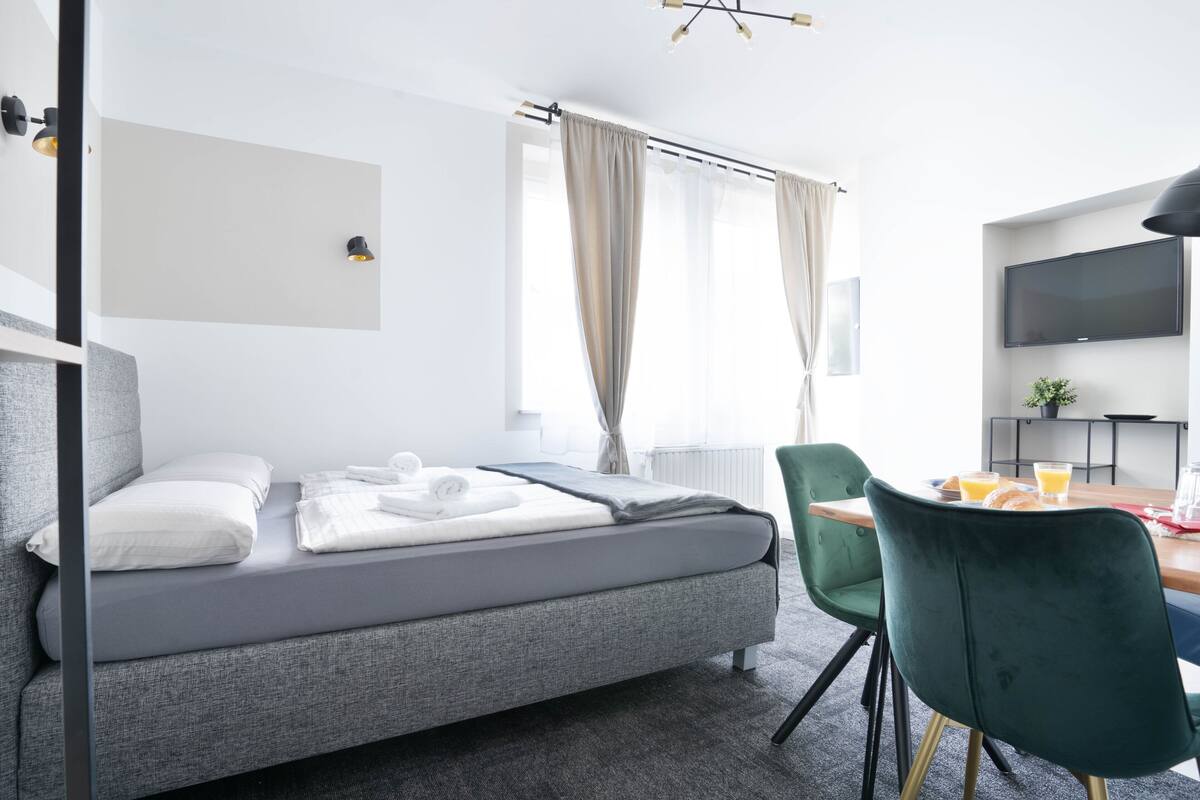 Een moderne hotelkamer met een grijze slaapbank, een eettafel met stoelen, een flatscreen televisie en een groot raam met gordijnen.