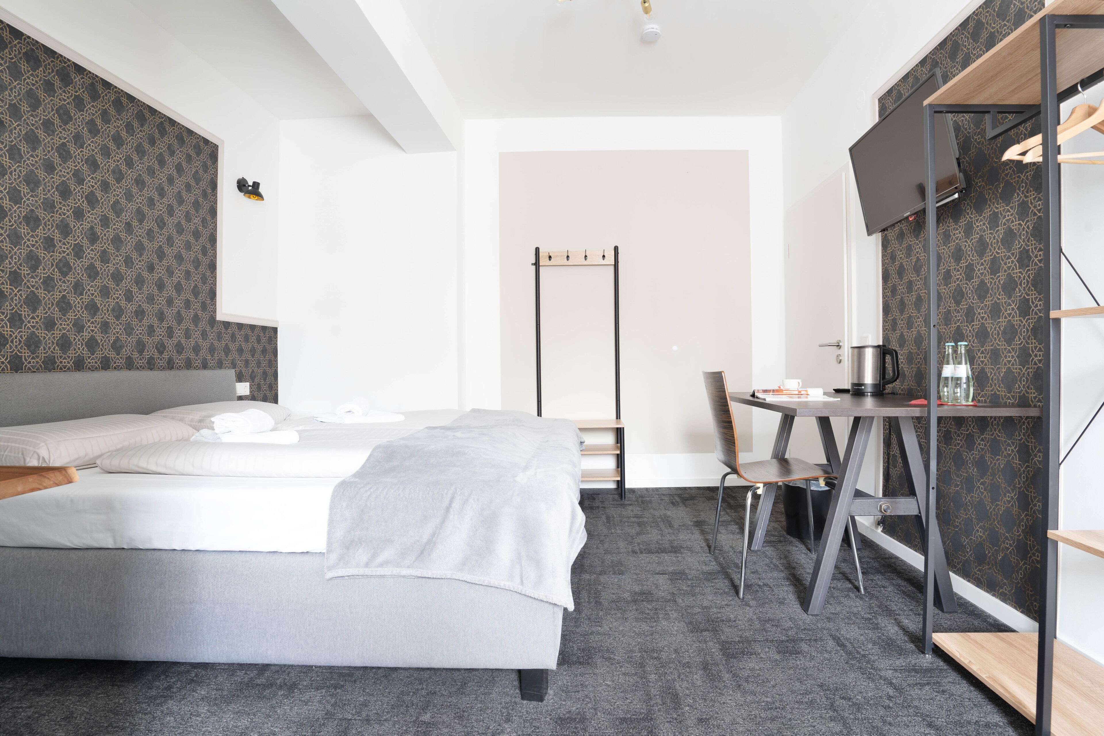 A modern hotel room with a bed, a desk with a chair, a TV, and a coat rack.