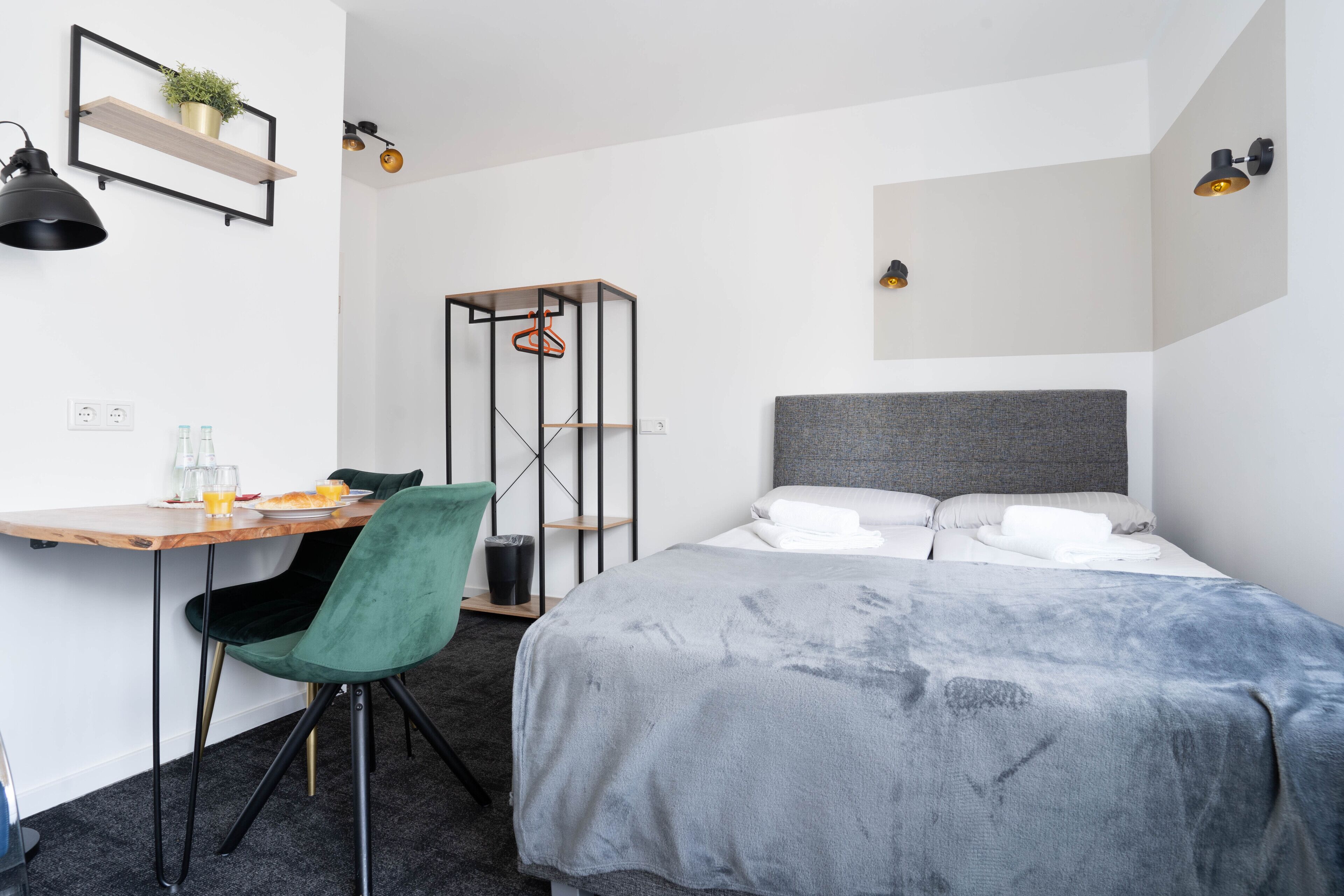 Et moderne hotelværelse med en seng, et skrivebord med stol, en lille hylde med en plante og et ophæng til tøj.