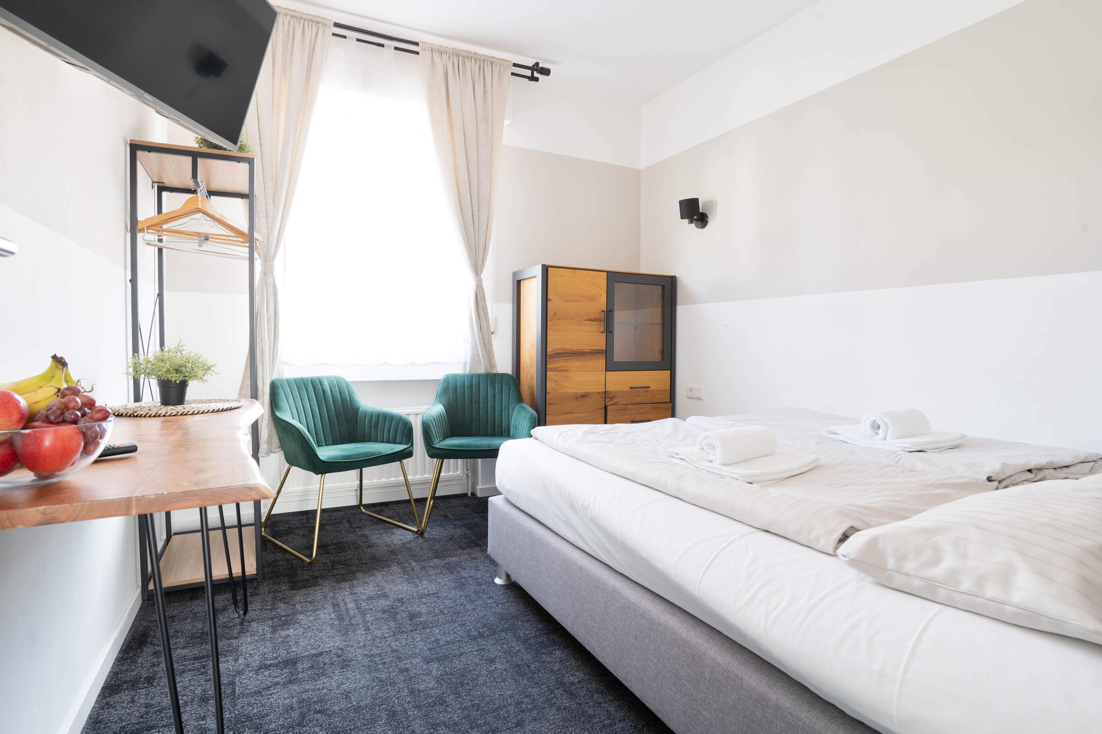 A hotel room with a bed, a desk with a chair, a small table with fruit, and a window with curtains.