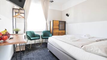 A hotel room with a bed, a desk with a chair, a small table with fruit, and a window with curtains.