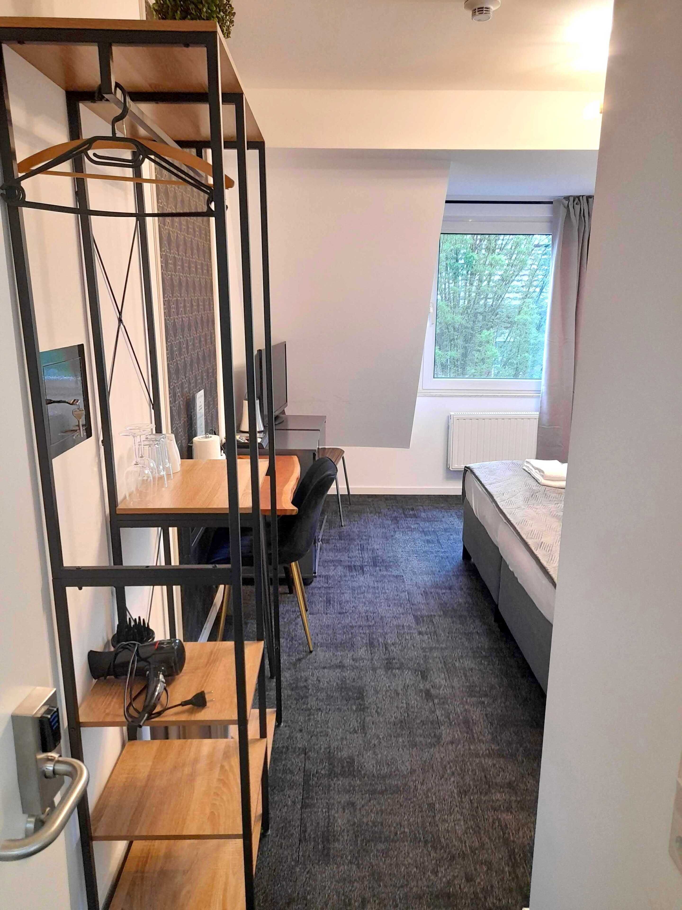Habitación de hotel compacta con literas, un escritorio con silla, un televisor pequeño y una ventana con vista a árboles.